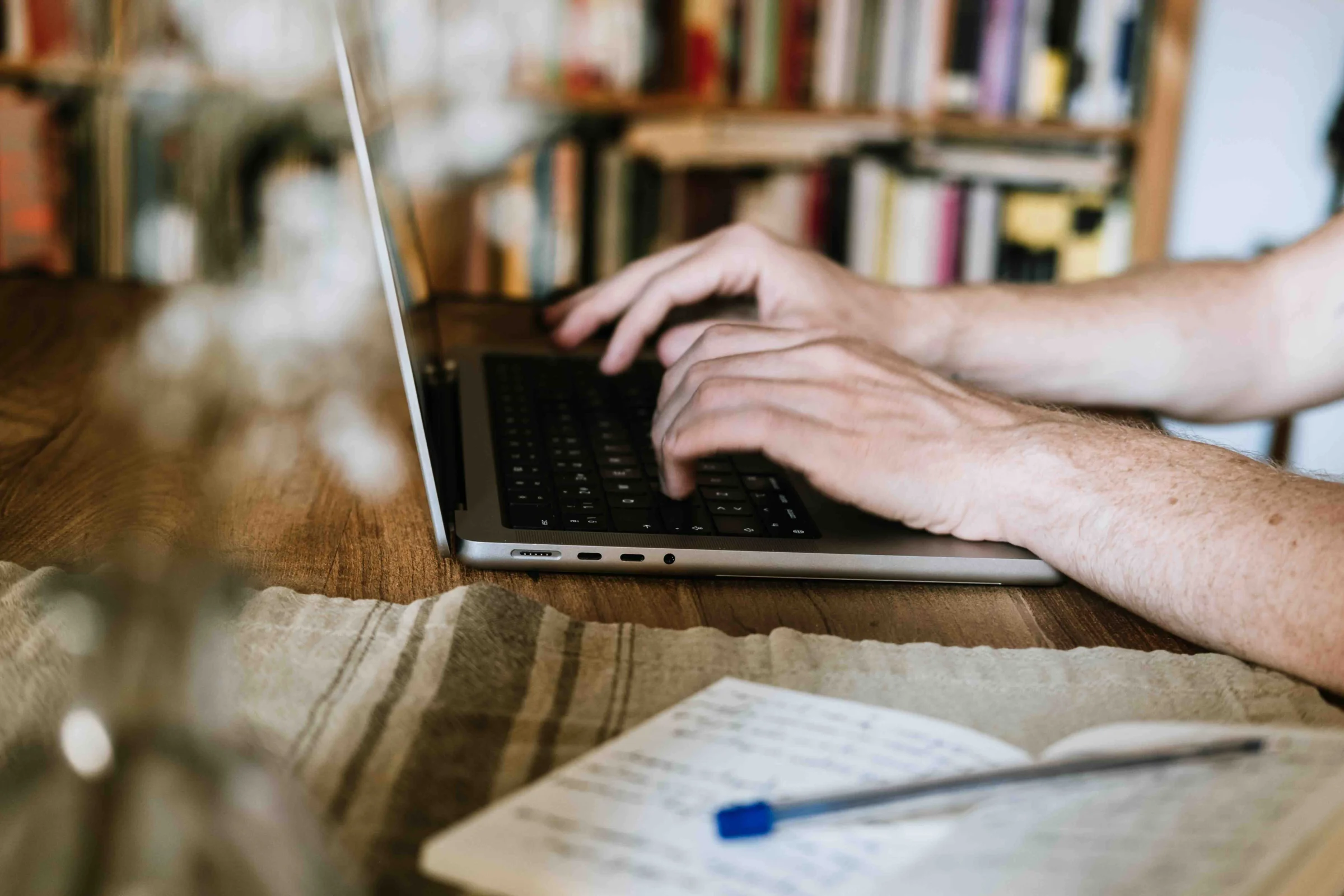 A person typing on a laptop with an open notebook and pen on a wooden desk lined with books.