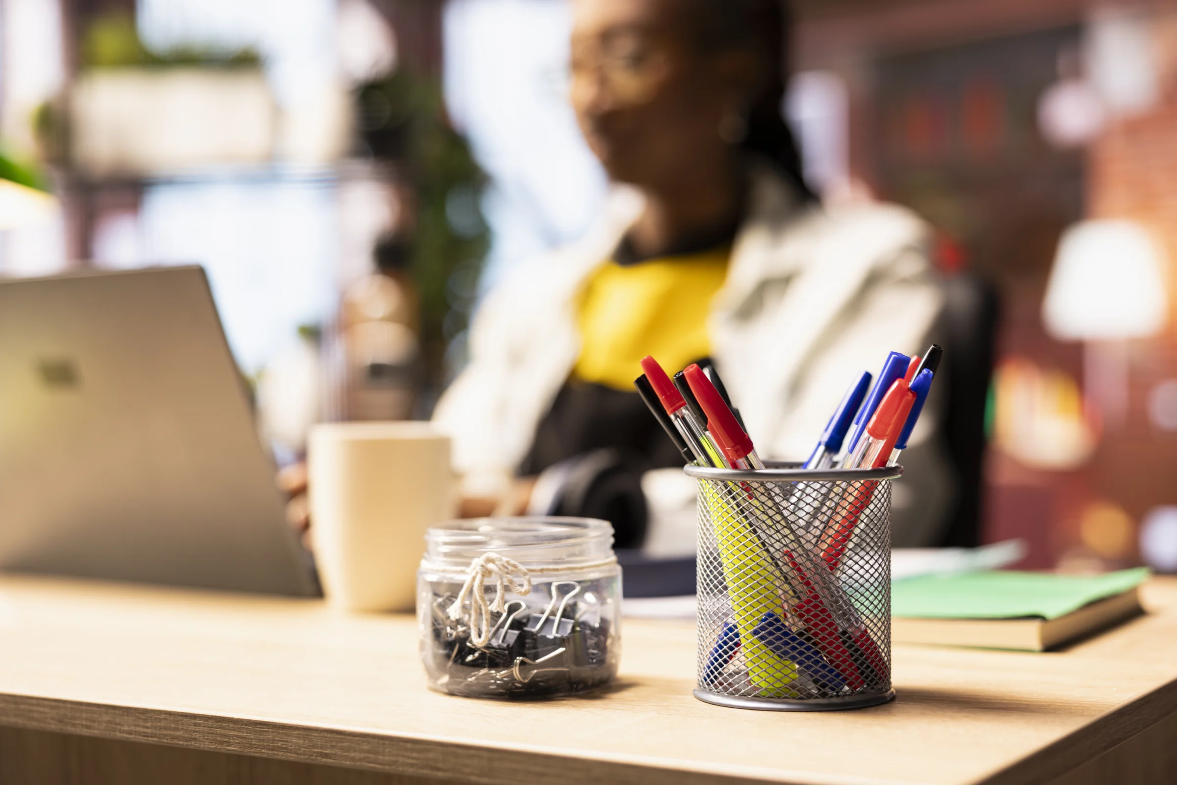A jar of pens and paper clips on a desk beside a laptop.