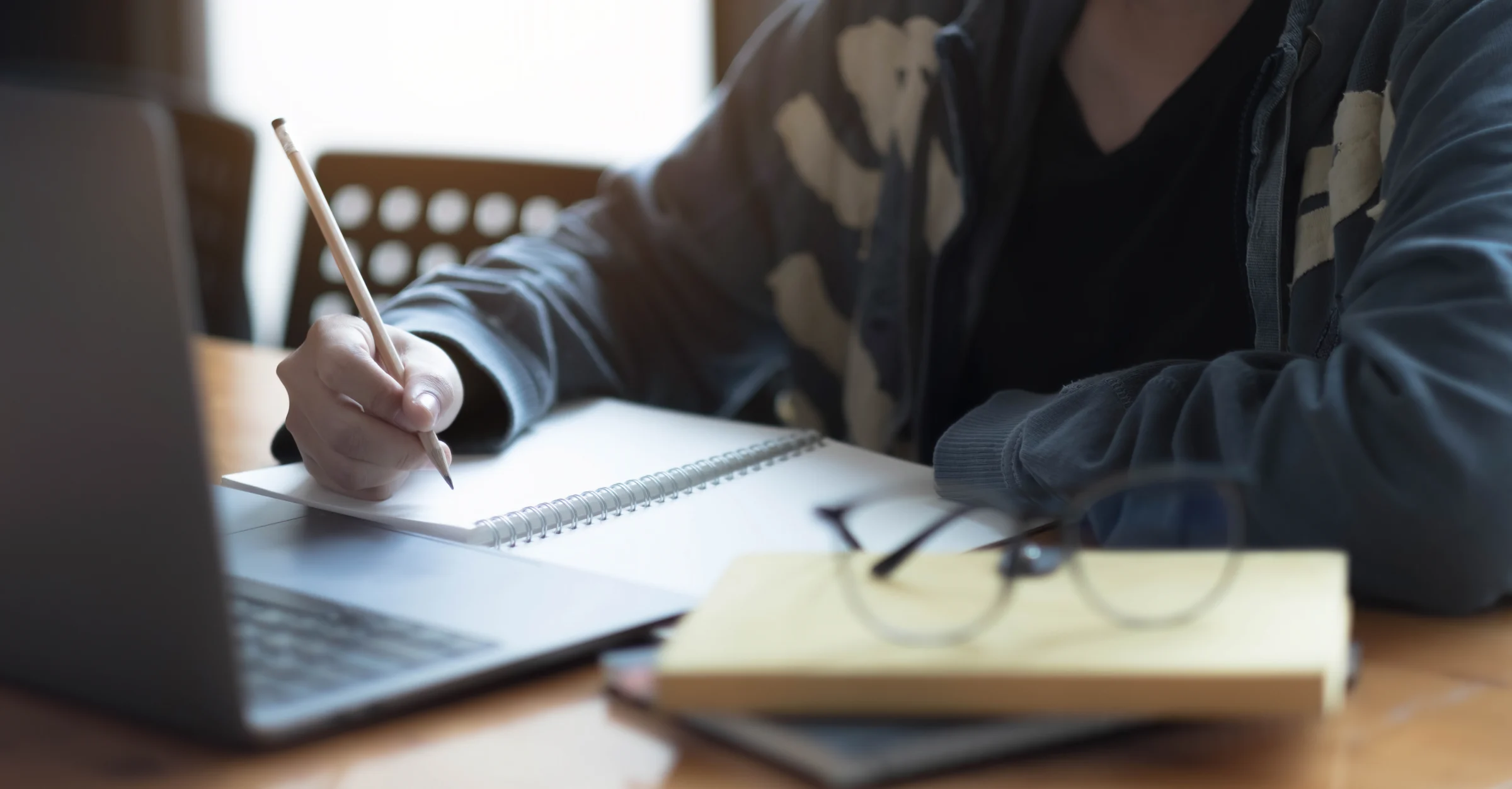 A hand writing notes in a spiral notebook beside an open laptop and a pair of glasses.